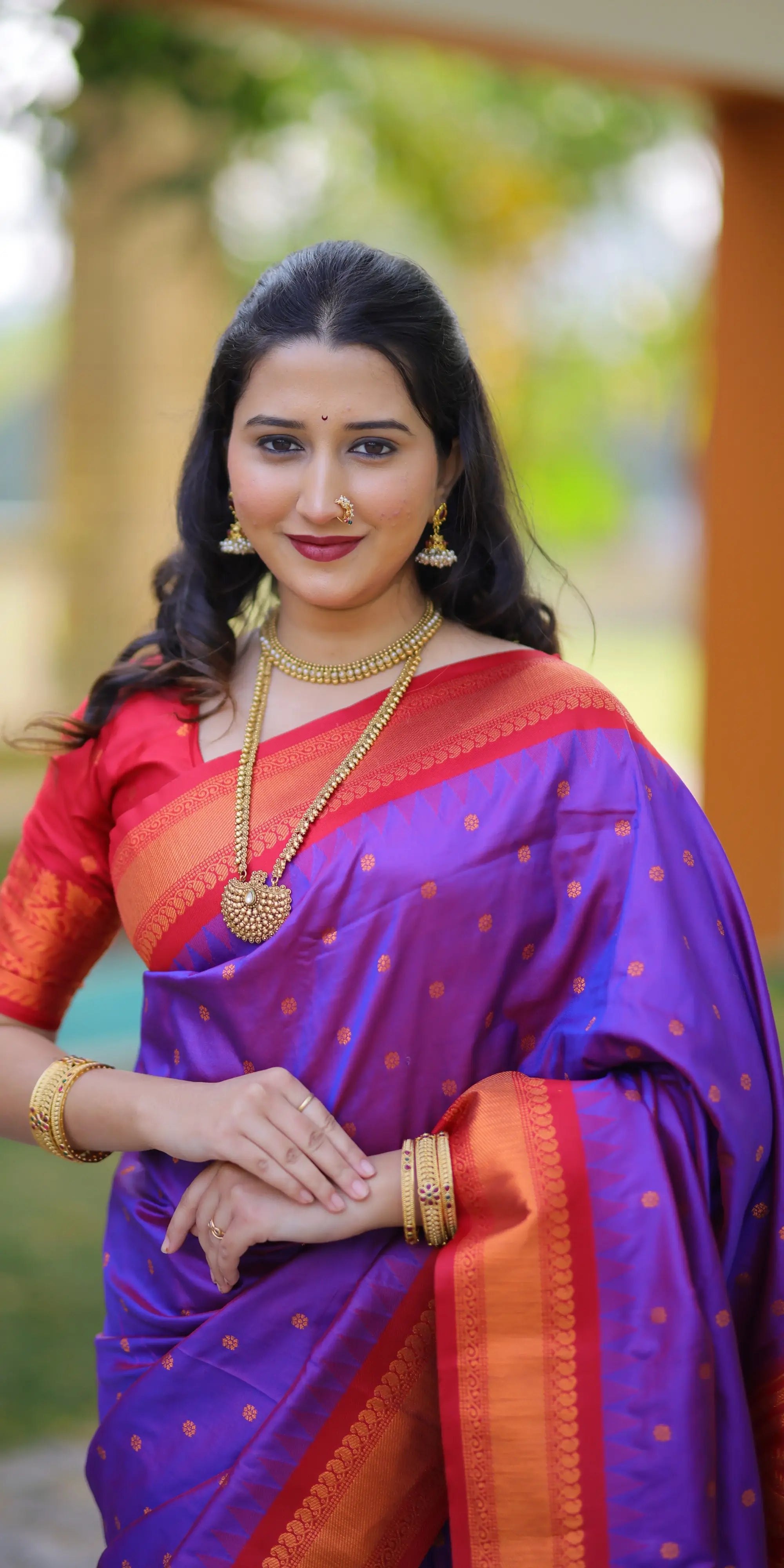 Woman wearing a traditional purple saree with a red blouse, standing outdoors.